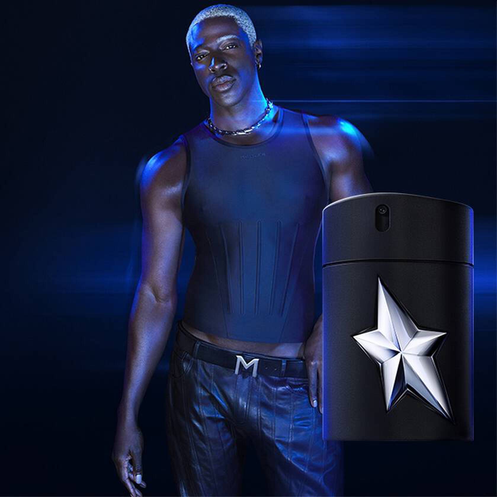 A fashionable man in a sleeveless top stands next to a sleek black fragrance bottle with a star logo, set against a dark, dynamic background.