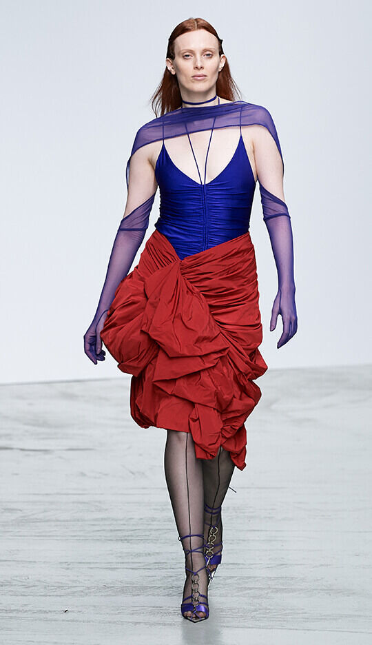 A model walks down the runway wearing a striking outfit featuring a deep blue top and a dramatic red skirt with ruffled detailing. She is accessorized with sheer purple gloves and a matching shawl. The setting is a minimalistic fashion show environment.