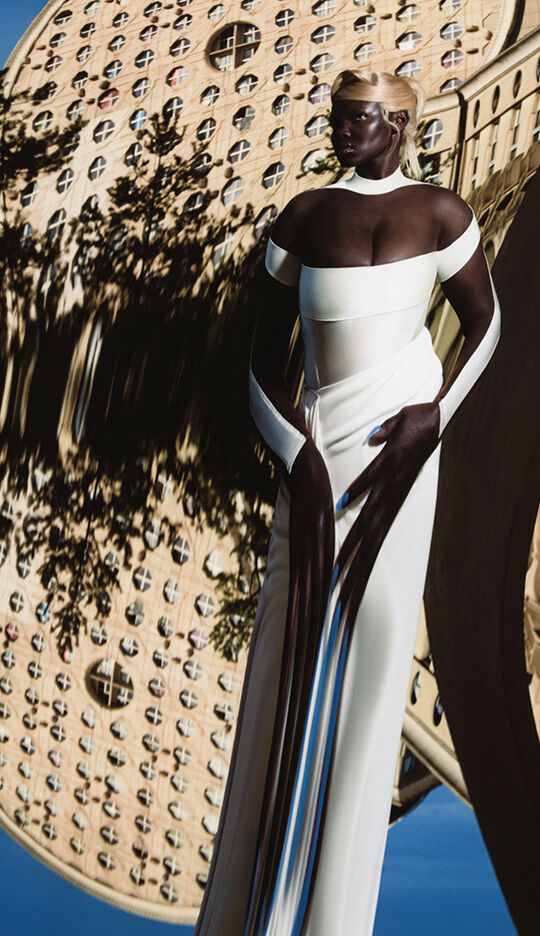 A model wearing an elegant white gown stands against a surreal, distorted background featuring a large, round architectural structure. The image has a dreamy, ethereal quality.