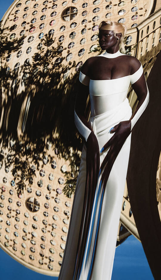 A model wearing an elegant white gown stands against a surreal, distorted background featuring a large, round architectural structure. The image has a dreamy, ethereal quality.
