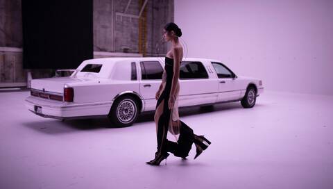 A model walks gracefully in a sleek black gown next to a white limousine in a minimalist studio setting.