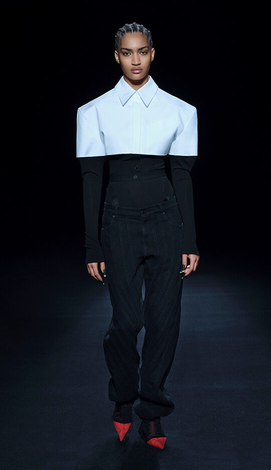 A model walks down a runway wearing a bold fashion look, featuring an oversized white collar shirt over a fitted black top and pinstripe pants, paired with striking red shoes.