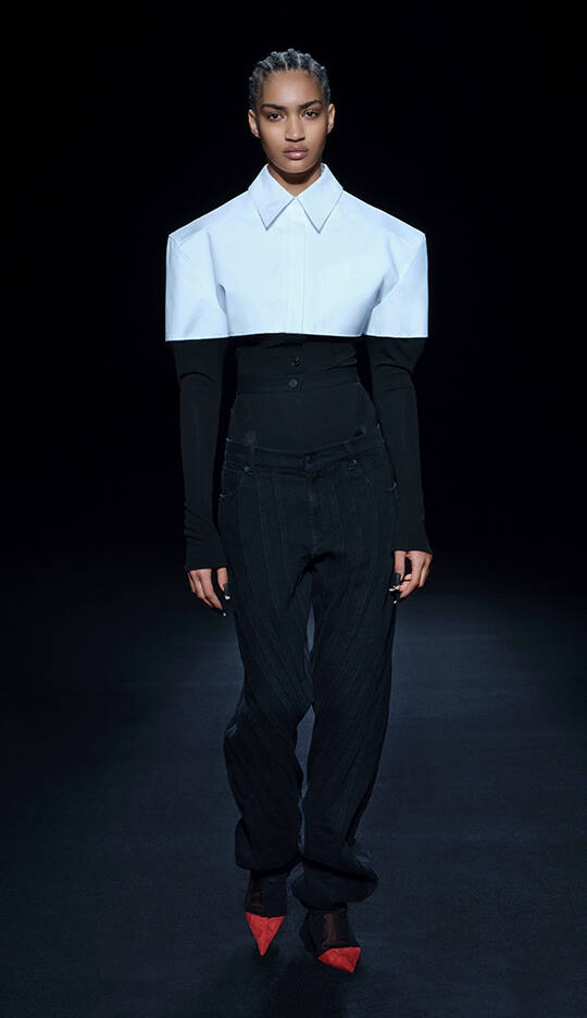 A model walks down a runway wearing a bold fashion look, featuring an oversized white collar shirt over a fitted black top and pinstripe pants, paired with striking red shoes.