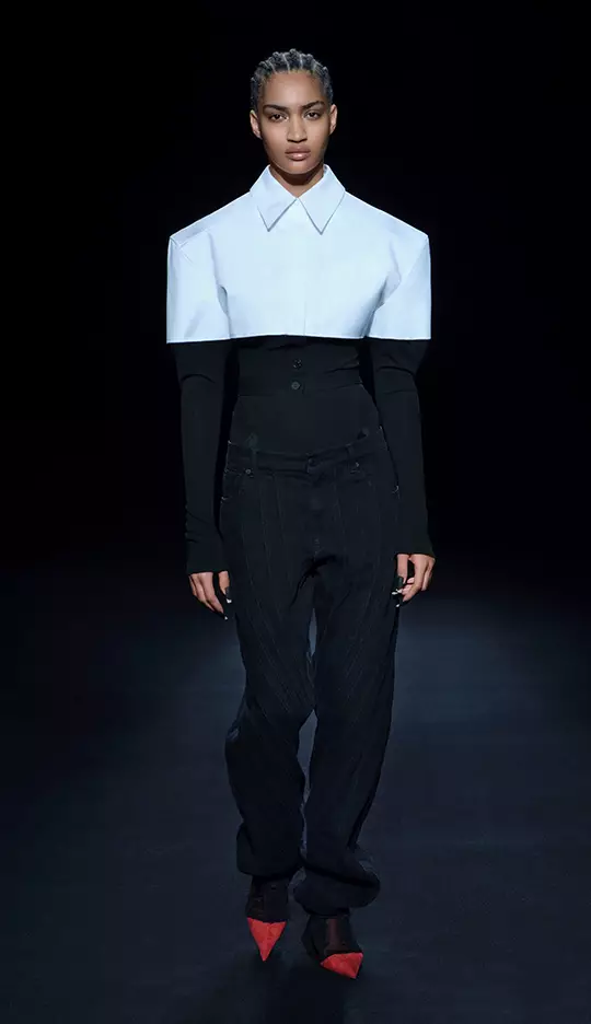 A model walks down a runway wearing a bold fashion look, featuring an oversized white collar shirt over a fitted black top and pinstripe pants, paired with striking red shoes.