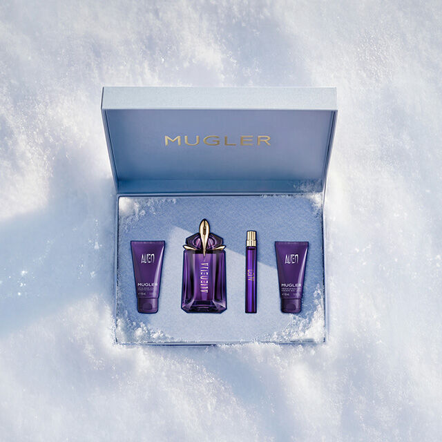 An open, textured ice-blue glistening Mugler gift set resting on snow, revealing an alien body lotion, shower gel, eau de parfum in 60ml as well as a travel sized 10ml. text reads "unforgettable gift sets"