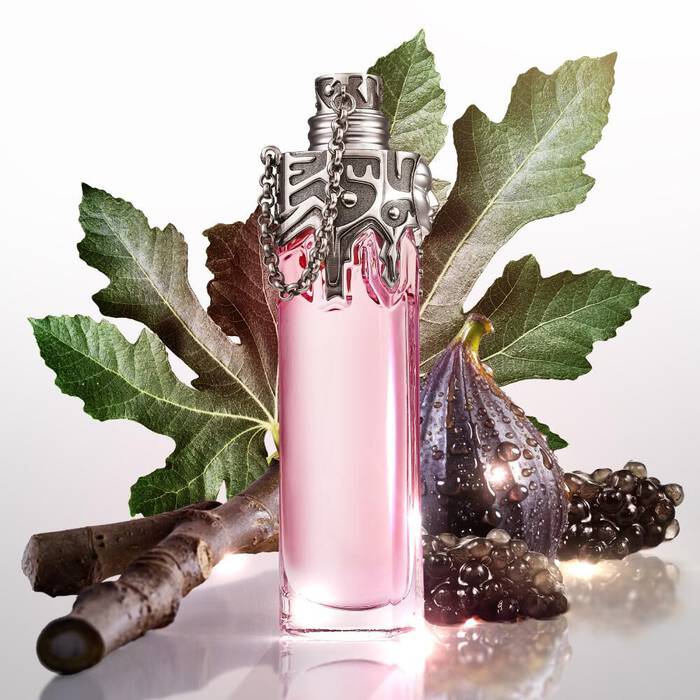 A rectangular pink perfume bottle with a detailed silver cap and chain, surrounded by large fig leaves, a fresh fig, dark berries, and a wooden branch, all set on a reflective surface.
