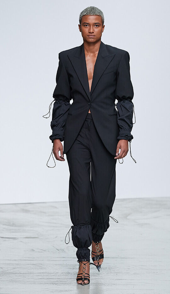 A model walks down the runway in a chic black pantsuit featuring a tailored jacket with puffed sleeves and fitted trousers with drawstring cuffs, complemented by strappy high-heeled shoes.