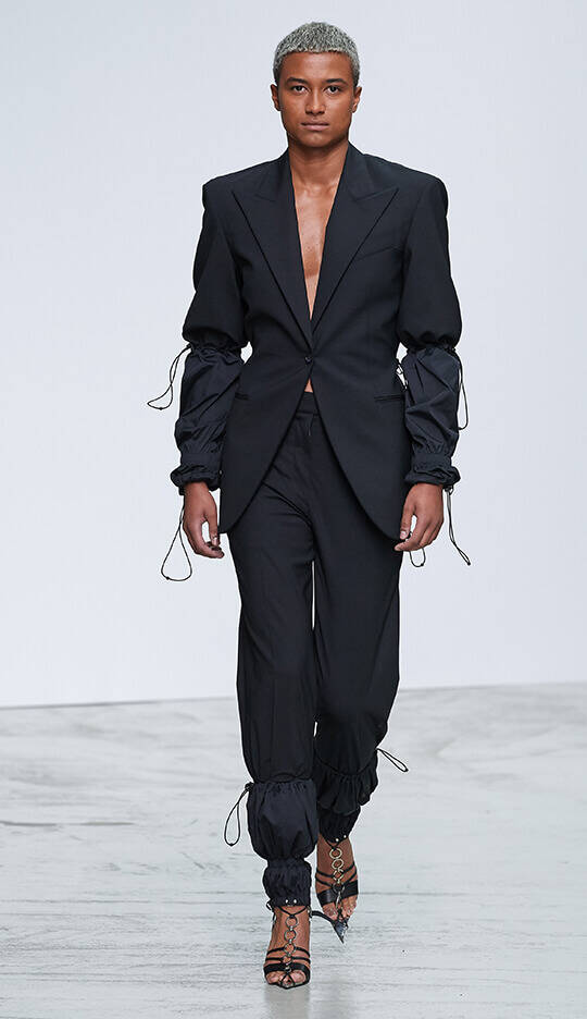 A model walks down the runway in a chic black pantsuit featuring a tailored jacket with puffed sleeves and fitted trousers with drawstring cuffs, complemented by strappy high-heeled shoes.