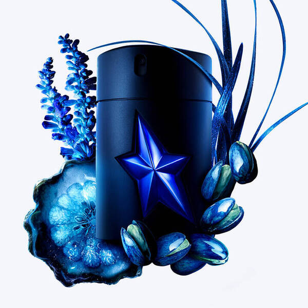 A matte black perfume bottle with a prominent blue star emblem, surrounded by vibrant blue lavender, a slice of blue agate, shiny blue-toned pistachios, and flowing blue grass-like elements, all set against a white background.