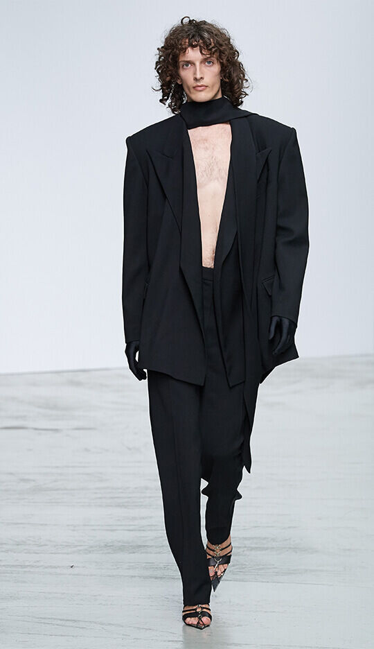 A model walks down the runway wearing a bold black oversized suit with a deep neckline and loose-fitting pants, accessorized with black gloves and strappy high-heeled shoes. The backdrop is a minimalist white setting.
