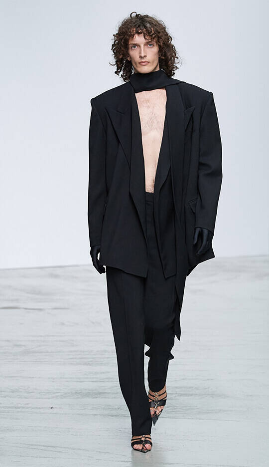 A model walks down the runway wearing a bold black oversized suit with a deep neckline and loose-fitting pants, accessorized with black gloves and strappy high-heeled shoes. The backdrop is a minimalist white setting.