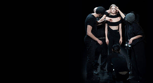 A dramatic performance scene with four figures in black clothing, two of whom are adjusting the top of a central figure, who is wearing a cropped top. The background is dark, creating a stark contrast with the performers.