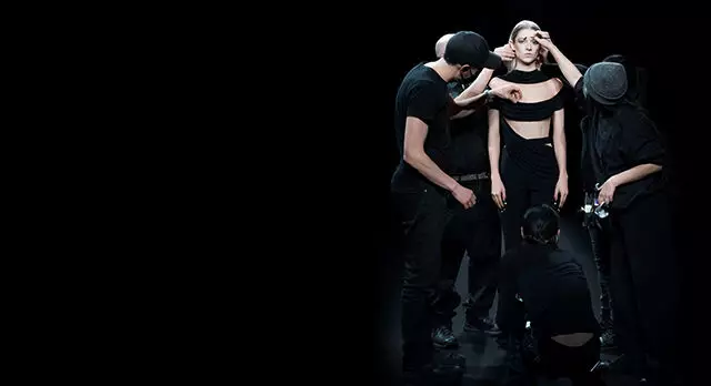 A dramatic performance scene with four figures in black clothing, two of whom are adjusting the top of a central figure, who is wearing a cropped top. The background is dark, creating a stark contrast with the performers.