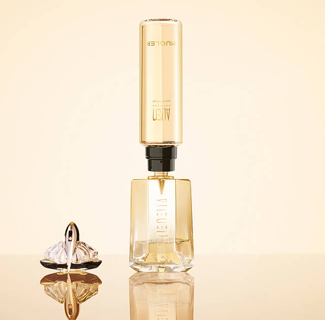 A stylish perfume bottle with a sleek, elongated design stands upright on a reflective surface, accompanied by a decorative crystal ornament. The bottle has a gold and clear exterior, exuding a luxurious aesthetic.