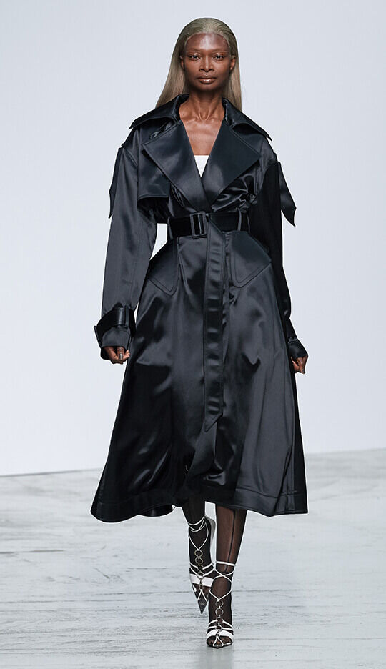 A model walks down the runway in a flowing black satin trench coat, styled with a cinched waist and a sleek silhouette, complemented by strappy heels.