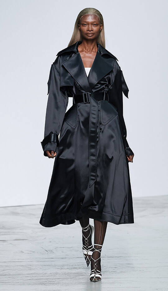 A model walks down the runway in a flowing black satin trench coat, styled with a cinched waist and a sleek silhouette, complemented by strappy heels.
