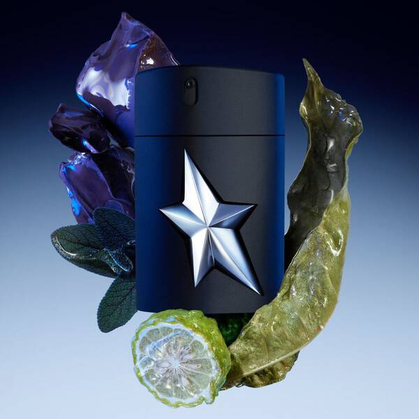 A black perfume bottle with a shiny silver star emblem, surrounded by glowing purple crystals, green leaves, a sliced bergamot, and a dried green leaf, set against a blue gradient background.