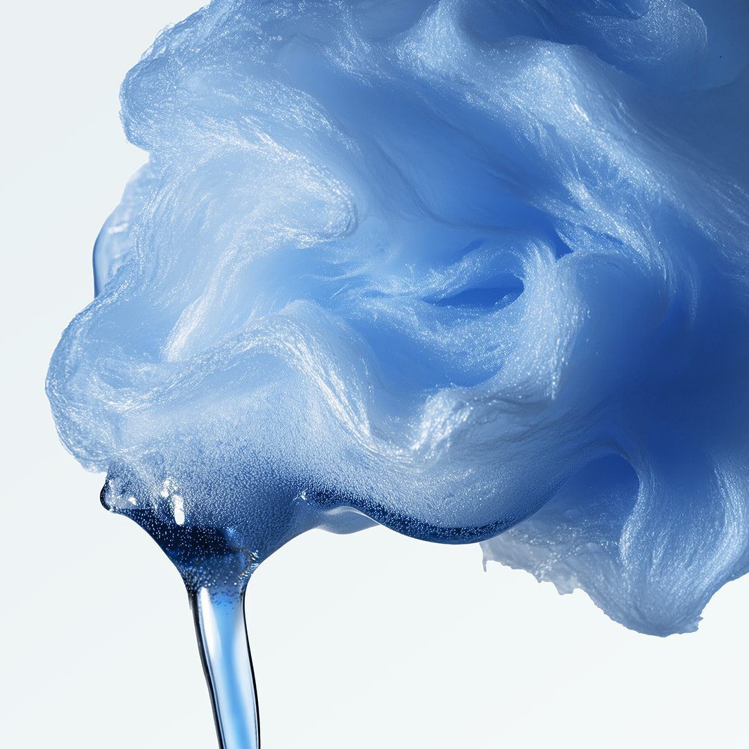 A close-up of a light blue, fluffy, and textured substance, resembling cotton candy or a sponge, from which a clear blue liquid drips down.