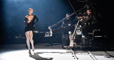 A performer in a stylish black dress poses on stage while a cameraman operates a large camera on a rig. The setting is dimly lit, creating an atmospheric backdrop.