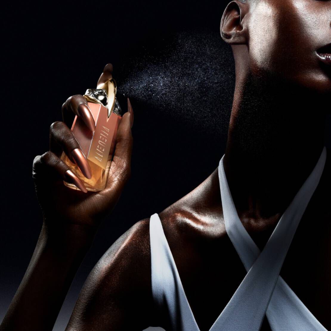 A close-up of a person holding a luxurious perfume bottle and spraying the fragrance, with a focus on their glistening skin and elegant outfit against a dark background.