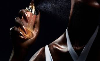 A close-up of a person holding a luxurious perfume bottle and spraying the fragrance, with a focus on their glistening skin and elegant outfit against a dark background.