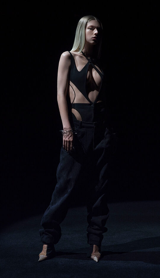 A model stands in a dark setting, wearing an edgy black outfit featuring a cut-out design and loose black pants. Her long straight hair and striking pose enhance the dramatic atmosphere.