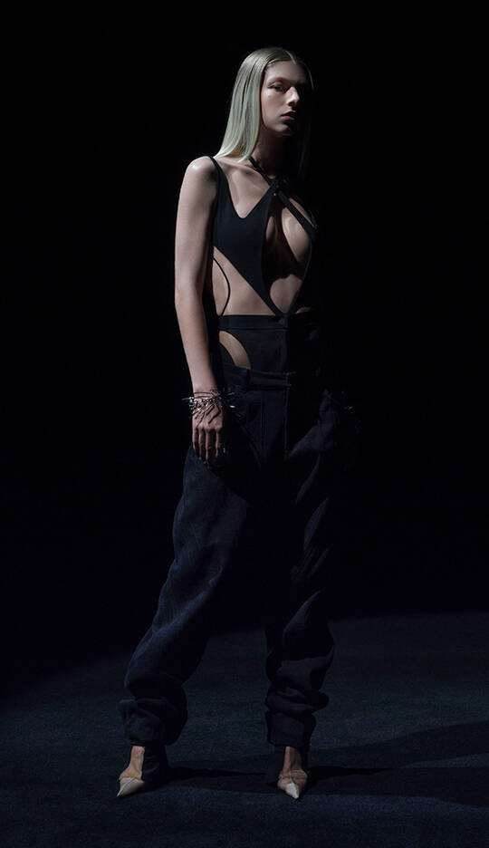 A model stands in a dark setting, wearing an edgy black outfit featuring a cut-out design and loose black pants. Her long straight hair and striking pose enhance the dramatic atmosphere.