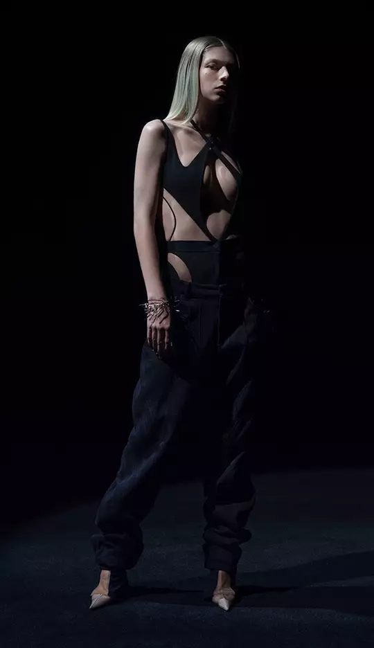 A model stands in a dark setting, wearing an edgy black outfit featuring a cut-out design and loose black pants. Her long straight hair and striking pose enhance the dramatic atmosphere.