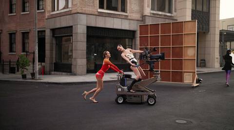 A scene on a film set featuring two individuals: one in a colorful red and orange outfit pulls a camera dolly, while another, partially clad, sits on the dolly, both appearing animated in a lively moment. Structures and a backdrop are visible in the urban setting.