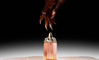 A close-up of a hand with long, elegant nails reaching towards a stylish perfume bottle on a marble surface, set against a dark background.