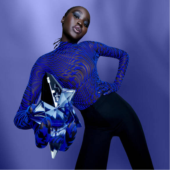 A model poses confidently against a purple background, wearing a blue patterned top and black pants. She holds a large, star-shaped bottle, suggesting a fragrance, and has bold makeup that enhances her striking look.