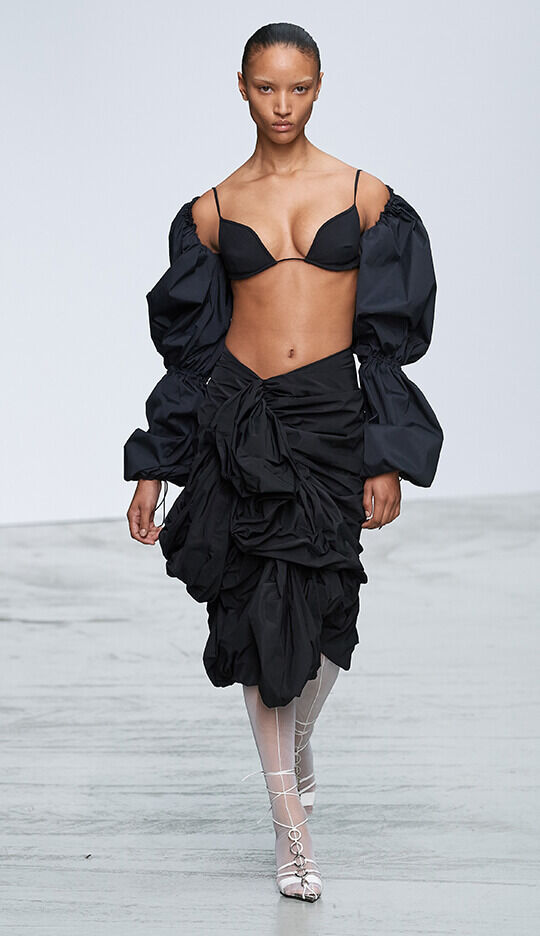A model walks down the runway wearing a black outfit with a fitted bra and a voluminous skirt adorned with ruffles, complemented by large, puffy sleeves.