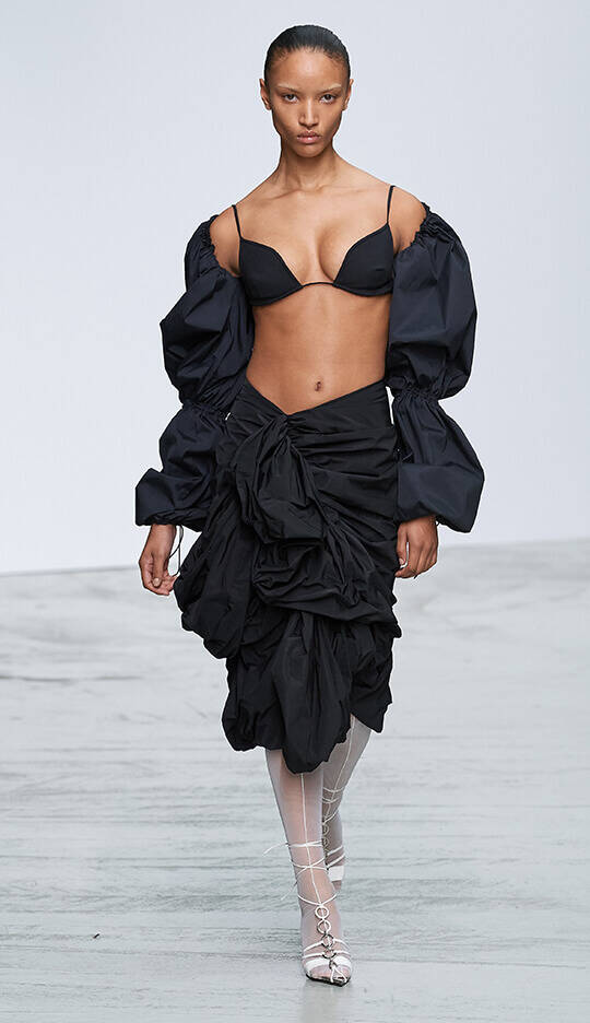 A model walks down the runway wearing a black outfit with a fitted bra and a voluminous skirt adorned with ruffles, complemented by large, puffy sleeves.