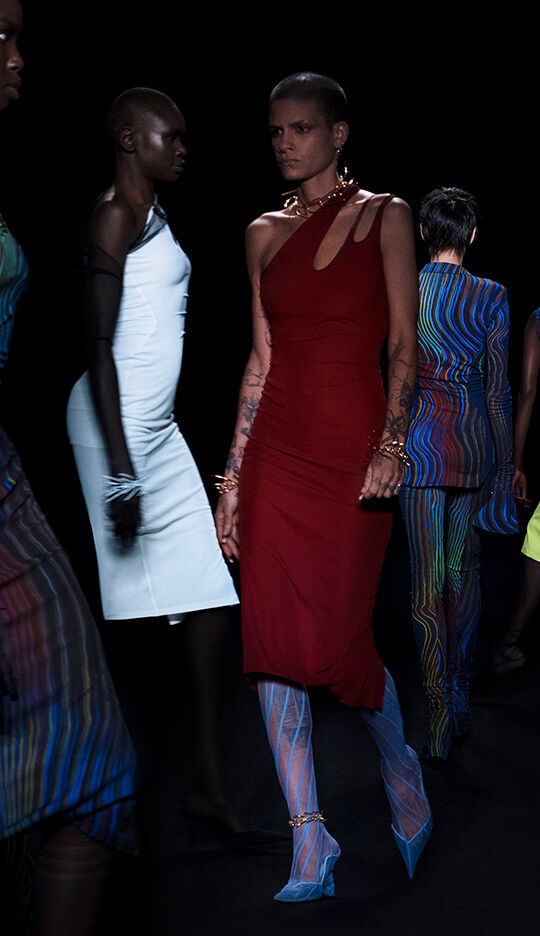 A group of models walks down a runway in stylish attire, showcasing a variety of colorful and uniquely designed outfits against a dark backdrop.