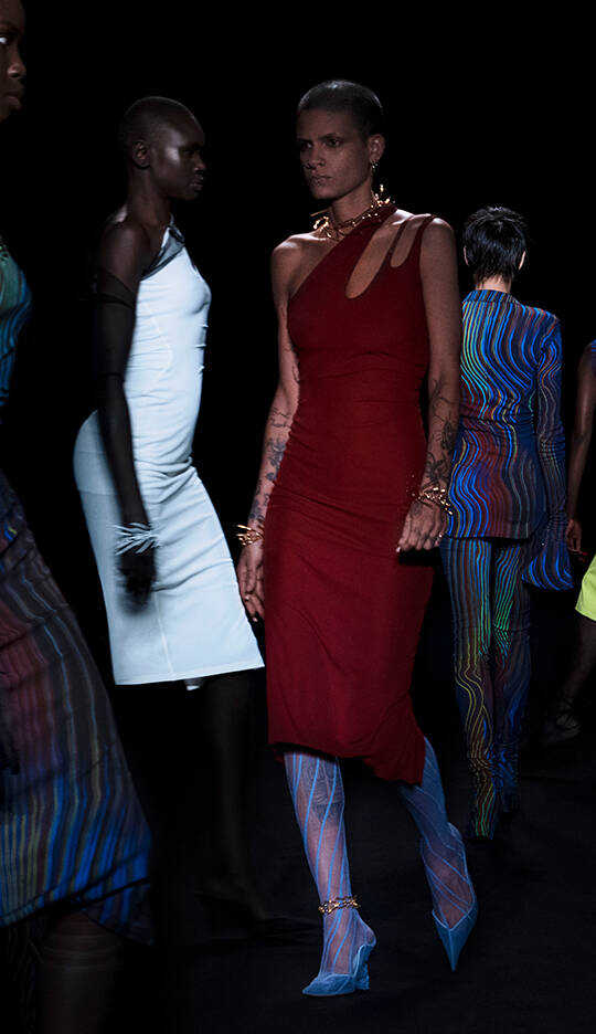 A group of models walks down a runway in stylish attire, showcasing a variety of colorful and uniquely designed outfits against a dark backdrop.
