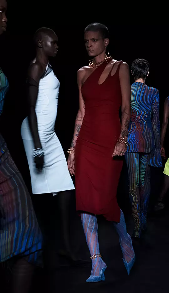 A group of models walks down a runway in stylish attire, showcasing a variety of colorful and uniquely designed outfits against a dark backdrop.