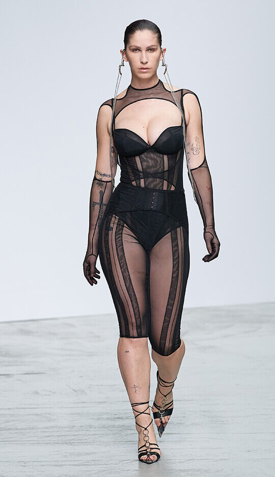 A model walks down the runway wearing a black sheer outfit with structured bodice and mesh details, complemented by long gloves and strappy heels, showcasing a bold fashion statement.