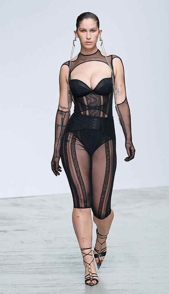 A model walks down the runway wearing a black sheer outfit with structured bodice and mesh details, complemented by long gloves and strappy heels, showcasing a bold fashion statement.