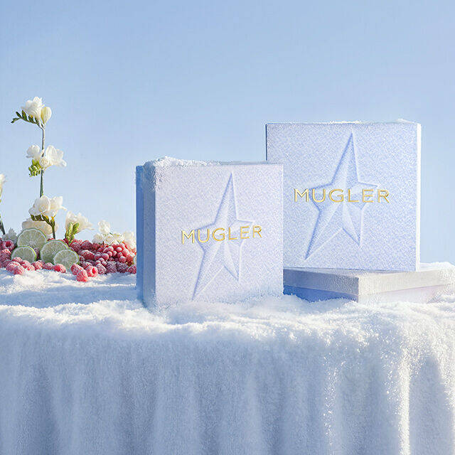 Two textured, ice-blue Mugler holiday gift sets featuring embossed star details, standing upright in a snowy winter landscape. text reads "the art of gifting. give a gift that makes an impression"