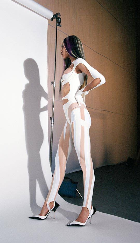 A model poses in a stylish, form-fitting outfit featuring cutouts and stripes, complemented by high-heeled shoes. The background is a simple studio setting with a soft light.