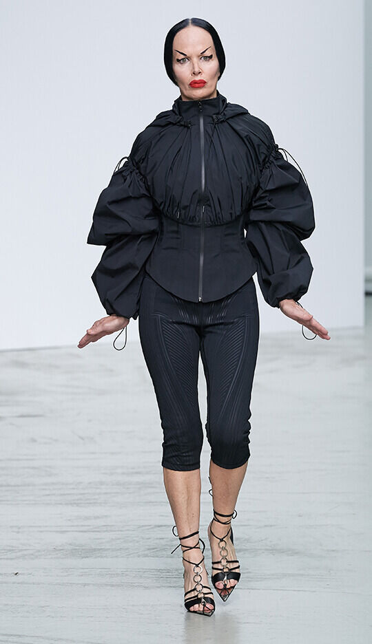 A model walks down the runway wearing a stylish black ensemble, featuring a structured jacket with oversized sleeves and form-fitting black leggings. The look is completed with strappy heels and a bold red lip.
