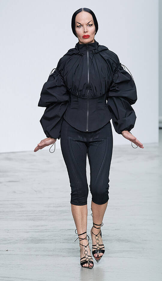 A model walks down the runway wearing a stylish black ensemble, featuring a structured jacket with oversized sleeves and form-fitting black leggings. The look is completed with strappy heels and a bold red lip.