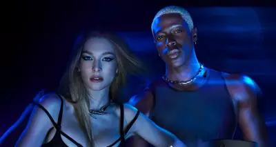 A stylish promotional image featuring a woman in a black outfit and a man in a tank top, both posing confidently against a dark background. They are surrounded by abstract light effects and a star-shaped product.