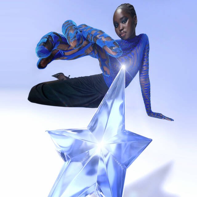 A model with dark skin and stylish attire poses against a blue background, reaching towards a large, translucent blue star.