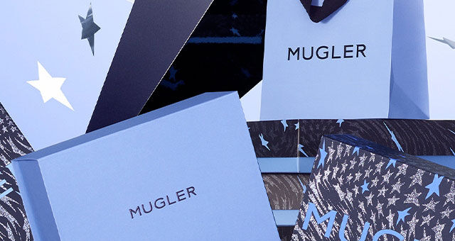 An artistic display of various Mugler gift boxes stacked and scattered against a soft blue background, with shiny stars floating around.