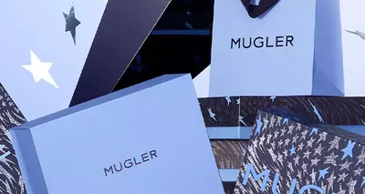 An artistic display of various Mugler gift boxes stacked and scattered against a soft blue background, with shiny stars floating around.