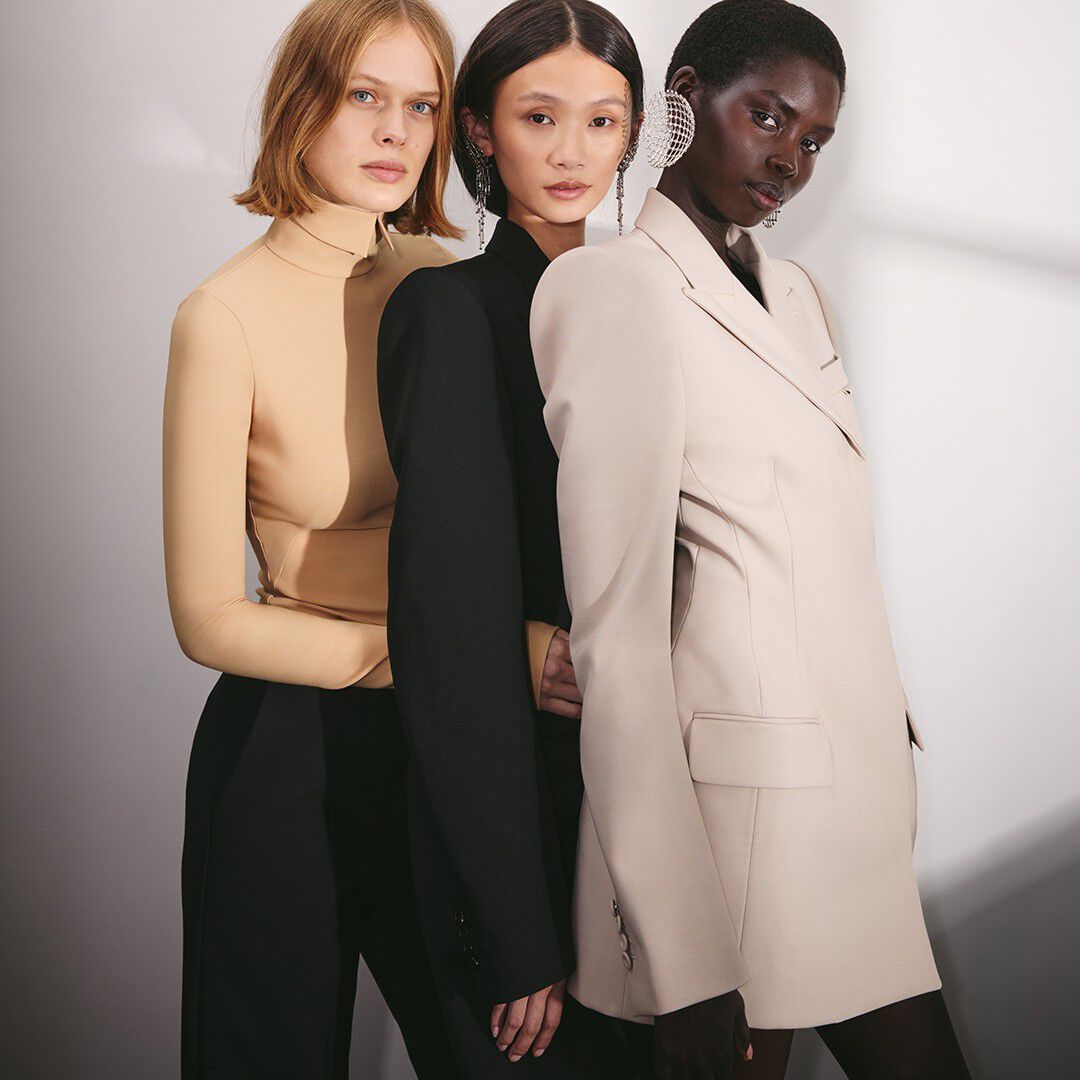 Three models standing together in a row against a neutral background, styled in elegant neutral-toned outfits including beige tops, a black blazer, and statement crystal earrings.