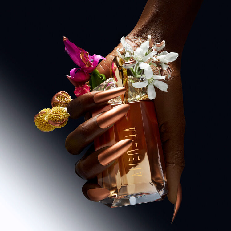 A hand with long, metallic nails holds a perfume bottle adorned with vibrant flowers, set against a gradient black background.