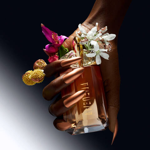 A hand with long, metallic nails holds a perfume bottle adorned with vibrant flowers, set against a gradient black background.