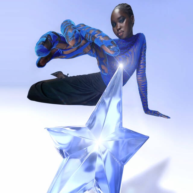 A model in a striking blue and black outfit poses with one arm extended towards a large, translucent blue star, set against a soft, light blue background.